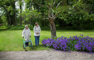 Bildet viser en eldre dame med rullator som er på tur i en hage med en yngre dame.
