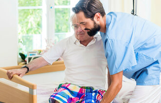 Bildet viser en pleier som løfter en eldre pasient ut av sengen. Begge er menn.