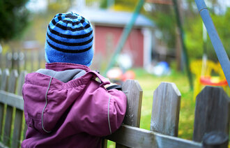 Bildet viser et barn som står med ryggen til mot et gjerde og ser lengselsfullt mot en lekeplass.