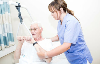 Bildet viser en sykepleier som hjelper en eldre mann med å sette seg opp i sengen