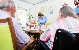 Bildet viser tre eldre personer som sitter rundt et bord på sykehjem. En sykepleier bøyer seg over den ene damen og smiler, mens en mannlig sykepleier står og smiler til de andre.