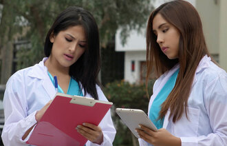 Bildet viser to kvinner med sykepleieruniform som står og snakker sammen utendørs. Den ene holder en mappe, mens den andre noterer på en skriveblokk.