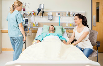 Bildet viser en pasient i sengen, mens en pårørende holder ham i hånda. Ved siden av står en sykepleier.