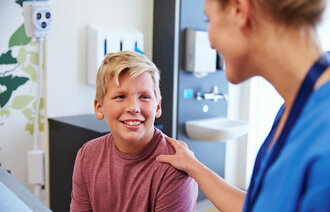 Bildet viser en ung gutt på et sykehusrom. En sykepleier legger hånden trøstende på skulderen til gutten. Gutten smiler.