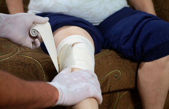 Bildet viser hendene til en sykepleier som bandasjerer et kne til en pasient hjemme i stolen sin.