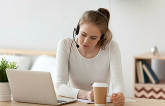 Bildet viser en ung kvinne som sitter i stua foran PC-en. Hun har hodetelefon med mikrofon på.
