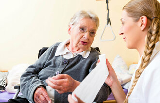 Bildet viser en eldre dame hjemme hos seg selv, og en sykepleier som ruller ut en bandasje mens hun snakker med pasienten. 