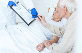 Bildet viser en syk kvinne i senga på sykehuset som har besøk av sin mann. En lege gir dem et skjema og en penn.