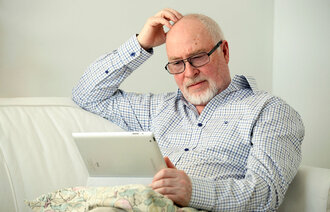 Bildet viser en eldre mann som sitter i en sofa med et nettbrett på fanget og klør seg litt i hodet