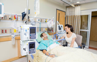 Bildet viser en mannlig pasient som ligger i sengen på sykehus. En kvinnelig pårørende sitter i en stol ved siden av.