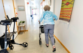 Eldre kvinne med gåstol i korridor fotografert bakfra.