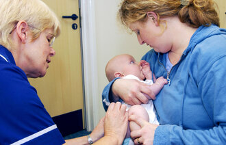 Bildet viser en baby som får vaksine av en helsesykepleier. Moren holder babyen trygt i armene og har blikkontakt med barnet