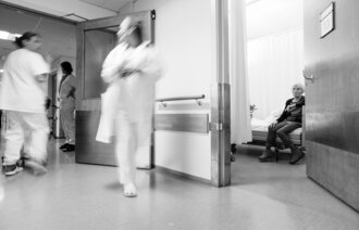 Bildet viser pasient i senga som venter på hjelp fra opptatt personale som fyker forbi.