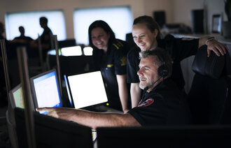 Bildet viser operasjonsleder 112, vaktleder 110 og operatør 113 som diskuterer foran en pc-skjerm.