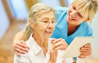 Bildet viser en eldre kvinner som ser på en iPad sammen med en sykepleier