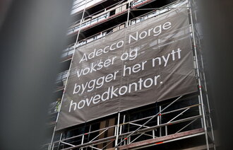 Bildet viser fasaden av et bygg under oppføring, med et banner der det står Adecco Norge vokser og bygger her nytt hovedkontor