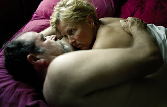 Et voksent par ligger i sengen halvveis under dyna og ser på hverandre