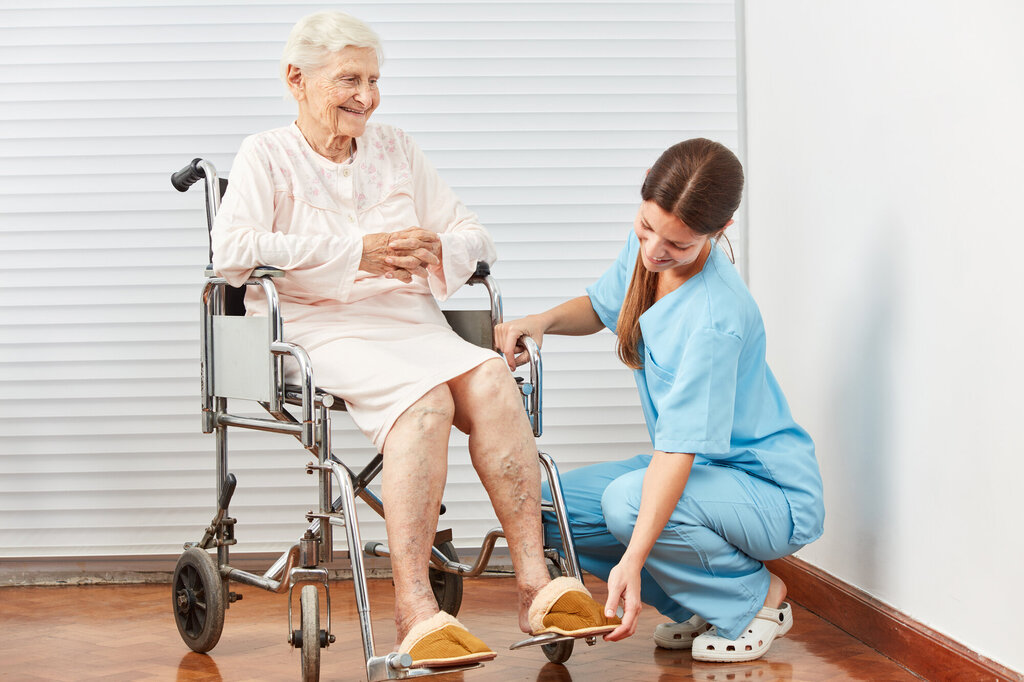 Bildet viser en sykepleier som sitter på golvet og hjelper en eldre dame i en rullestol.