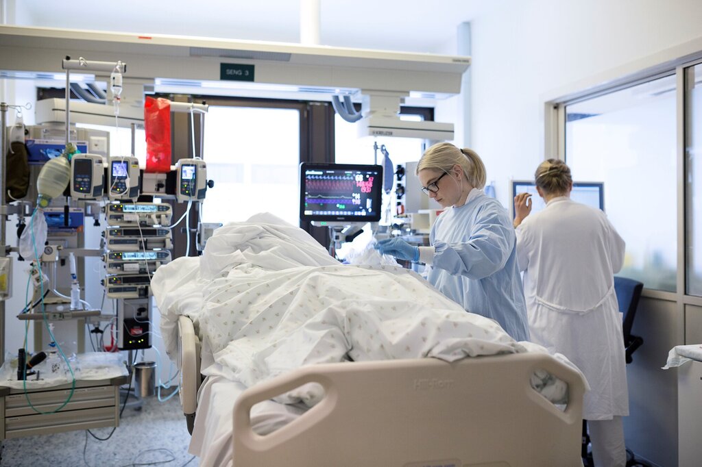Bildet viser en intensivavdeling, der en pasient ligger i sengen mens en sykepleier står ved siden av. I bakgrunnen står legen og sjekker på en skjerm