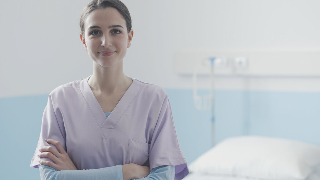 Bildet viser en ung sykepleier som står med armene i kors foran en sykehusseng