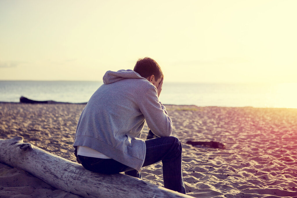 Bildet viser en ung mann som sitter på stranda med hodet i hendene