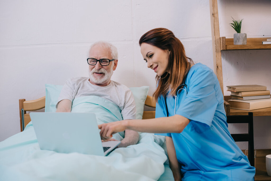 En sykepleier sitter ved sengen til en pasient. Han har en laptop i fanget, og sykepleieren peker på skjermen.