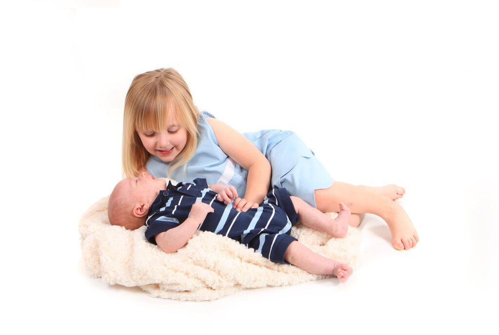 Bildet viser et spedbarn som ligger på ryggen på et skinn, og en litt eldre jente som ligger ved siden av og kikker ned på barnet