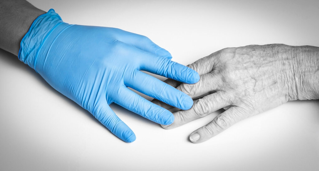 Bildet viser en hånd iført en plasthanske som berører hånden til et eldre menneske