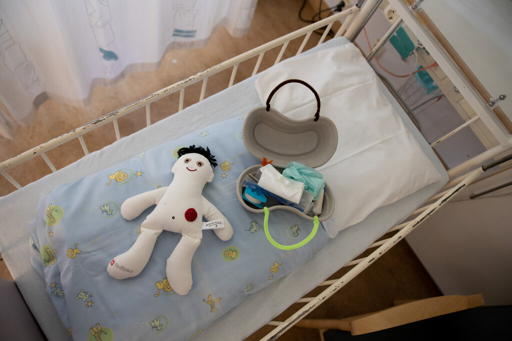 Bildet viser en dukke og et førstehjelpsskrin laget av et pussbekken, som ligger i en sykehusseng.
