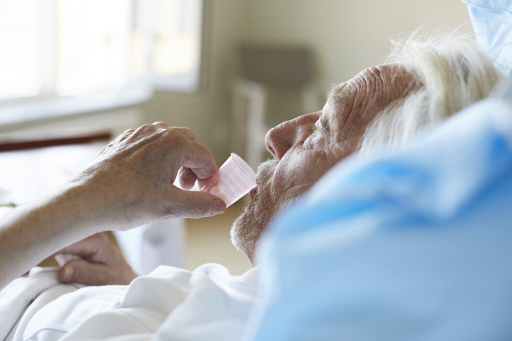Bildet viser en eldre, syk mann i senga som drikker medisin fra et plastglass