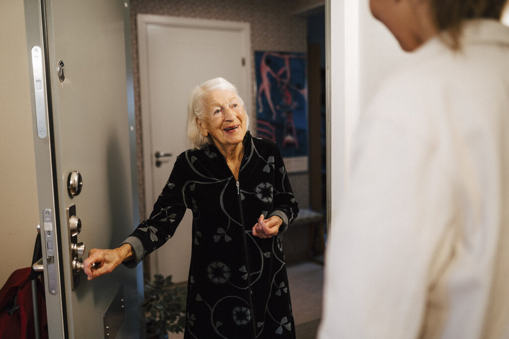 Bildet viser en smilende eldre kvinne som åpner døren for hjemmesykepleien