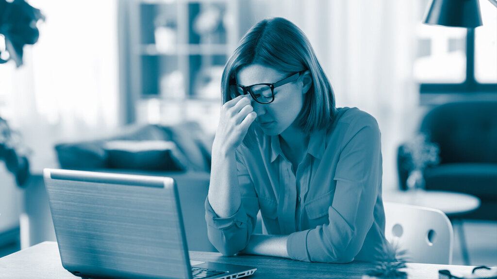 Bildet viser en dame som sitter hjemme foran en laptop. Hun ser sliten ut og tørker øynene under brillene