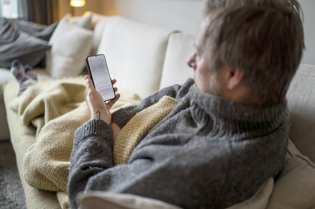 Bildet viser en mann som ligger under et pledd på sofaen og ser på mobilen