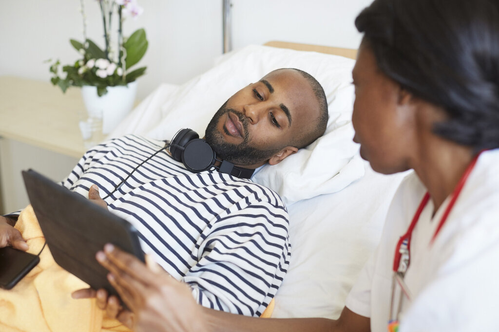Bildet viser en pasient som ligger i sengen og ser på en håndholdt skjerm, mens en sykepleier sitter på sengekanten