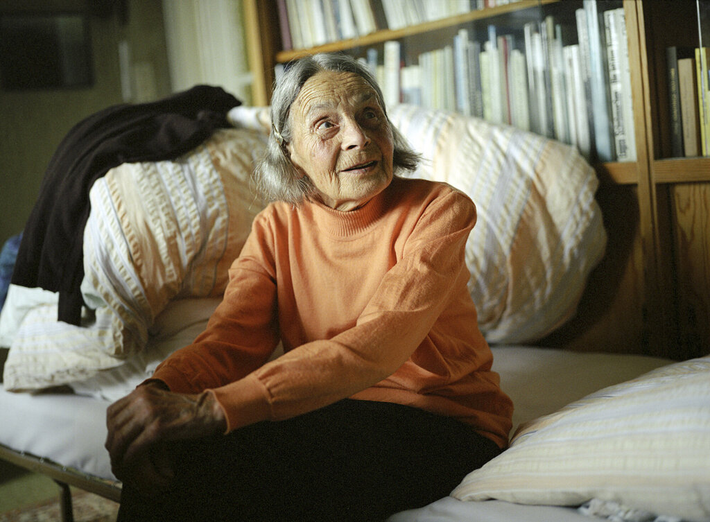 Bildet viser en eldre kvinne som sitter på en seng i hjemmet sitt