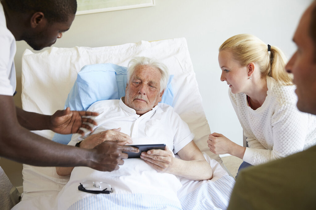 Bildet viser en syk, eldre mann i sengen. En sykepleier står over ham med et nettbrett. En ung dame er pårørende ved sengekanten
