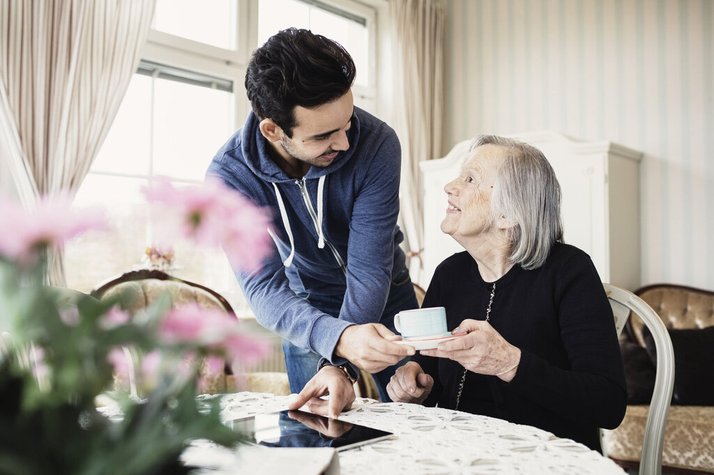 Bildet viser en pleier som gir en smilende, eldre pasient en kopp kaffe