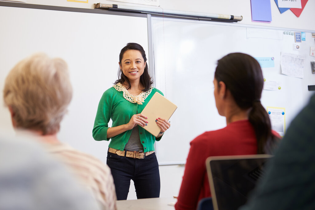 Bildet viser ei dame som står foran en whiteboard og underviser. Hun holder en skriveblokk i hånda. I rommet ser vi to voksne, sittende damer bakfra