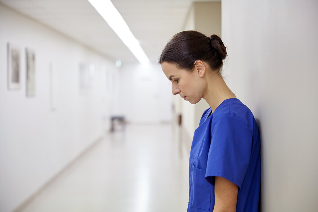 Bildet viser en sykepleier med bøyd hode som lener seg til en vegg. Hun er trist.