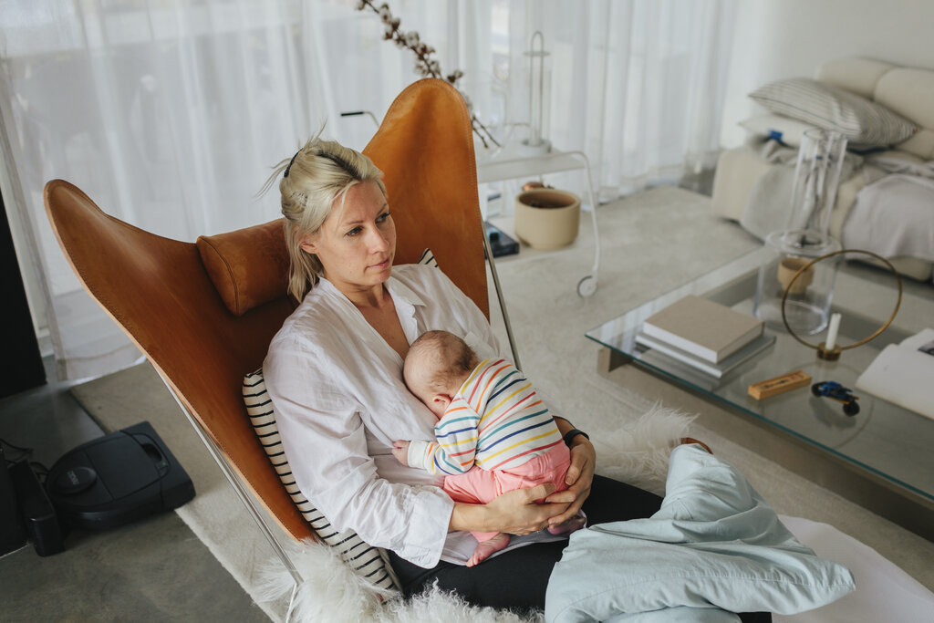 The photo shows a young mother sitting in a chair in her living room. She looks tired and holds her newborn baby in her lap.