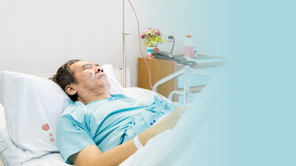 The photo shows a sick man lying in a hospital bed. On the bedside table there are flowers and a bottle. On the pillow there are some drops of blood