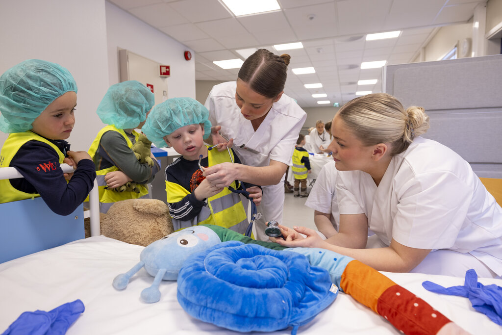 Bildet viser en gruppe med barn som står sammen med to sykepleierstudenter. De undersøker en kose-orm med et stetoskop.