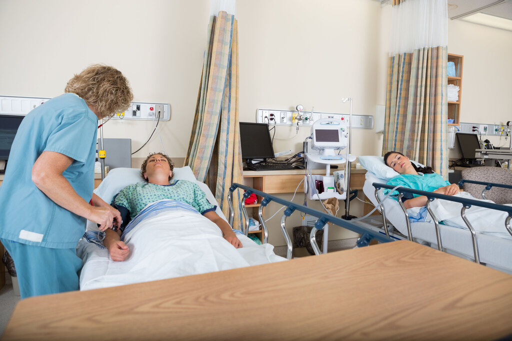 En sykepleier undersøker en pasient på sengepost. En annen pasient ligger i sengen ved siden av.