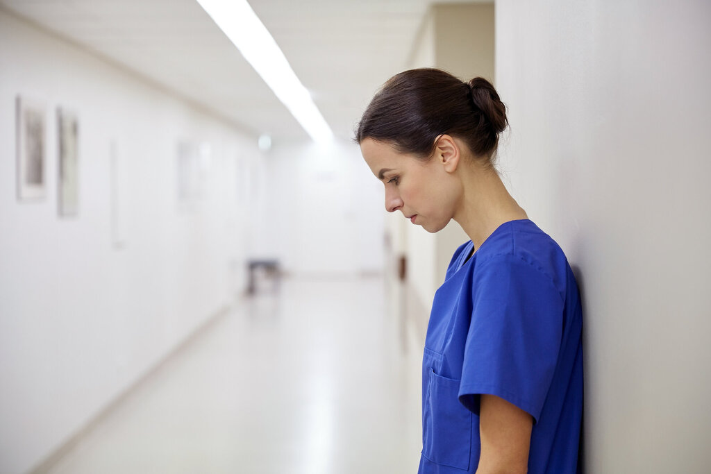 Kvinnelig helsearbeider står inntil veggen i en sykehuskorridor og kikker i golvet. Hun virker sliten eller fortvilet.