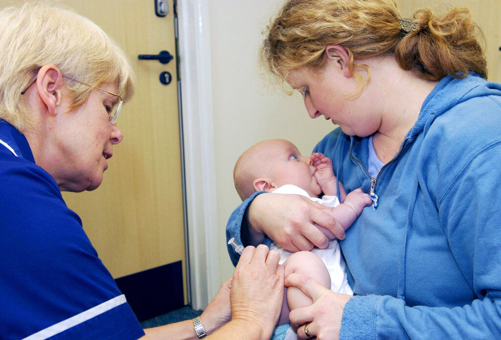 Bildet viser en baby som får vaksine av en helsesykepleier. Moren holder babyen trygt i armene og har blikkontakt med barnet