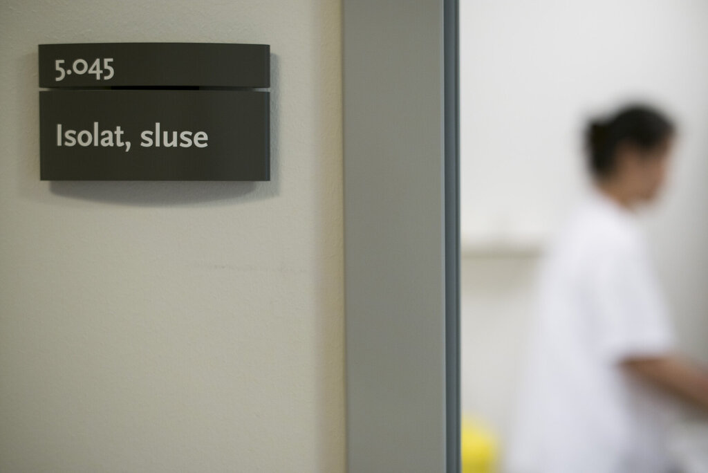Bildet viser et skilt der det står isolat, sluse og gjennom et vindu i døren skimtes en sykepleier