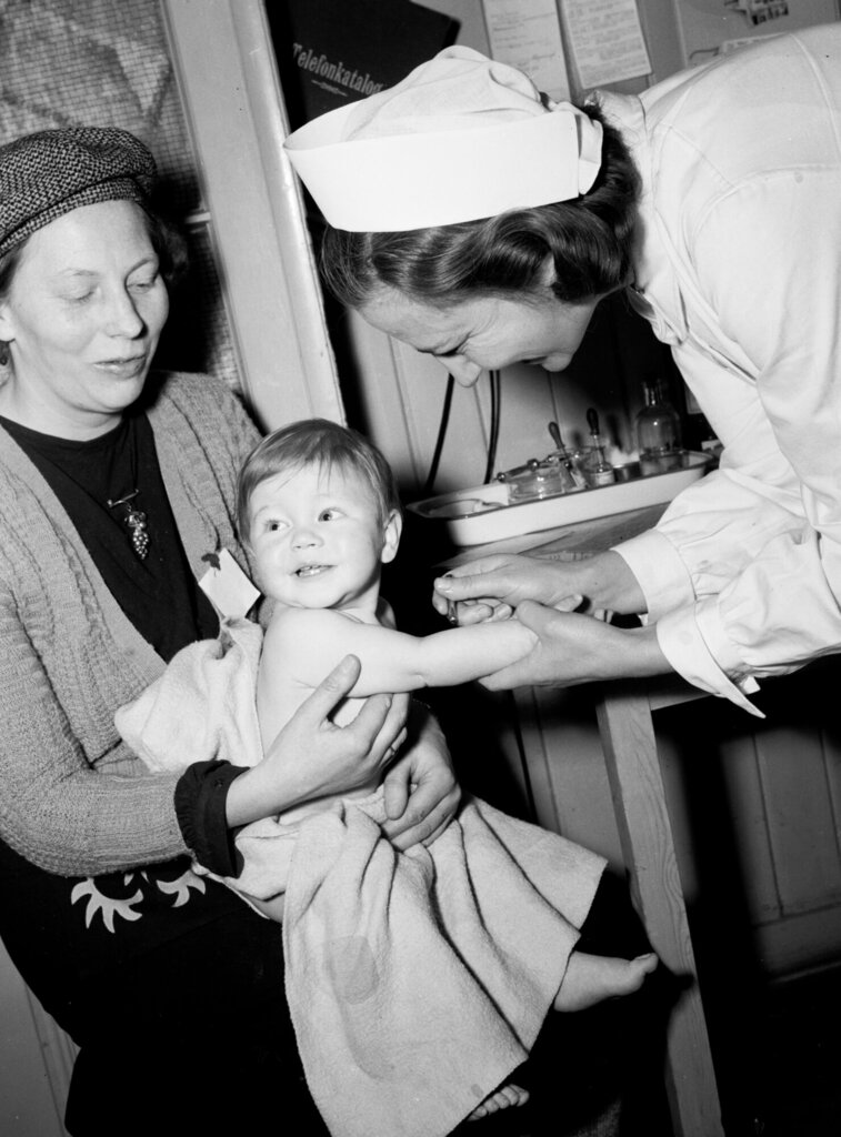 Bildet viser en kvinne som har barnet sitt på fanget og en helsesøster som er i ferd med å sette en vaksine.