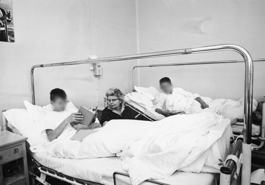 Bildet viser to gutter i hver sin sykehusseng, en eldre kvinne sitter mellom dem.