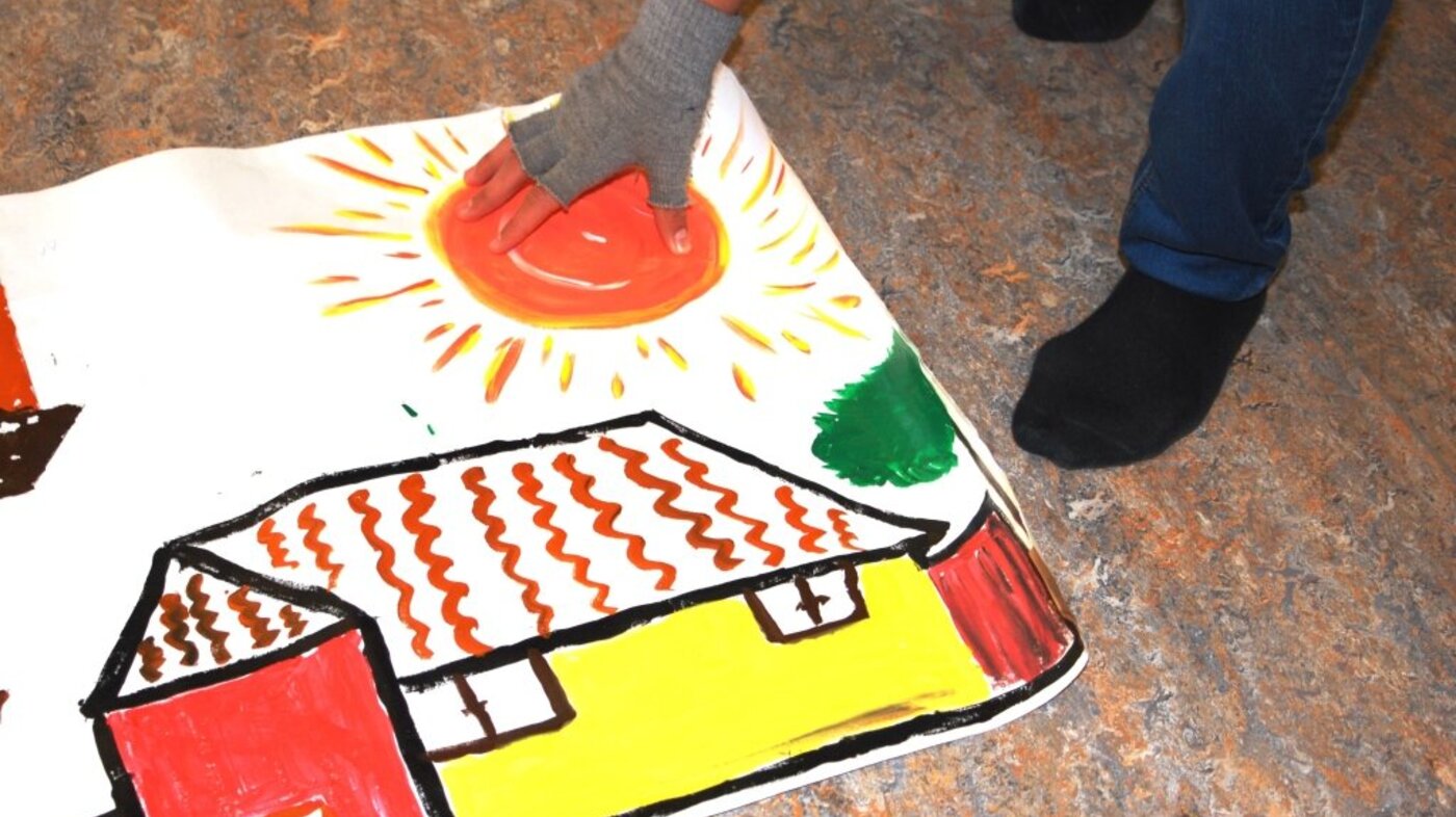 Bildet viser et maleri av et hus og sol, og en hånd lagt over sola.