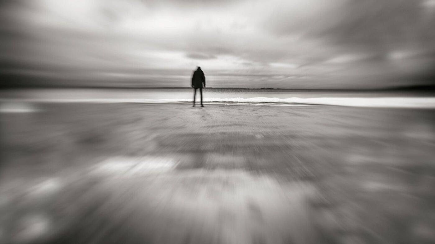 Abstrakt sort-hvitt bilde av en skikkelse som står på en strand og ser ut over havet. 